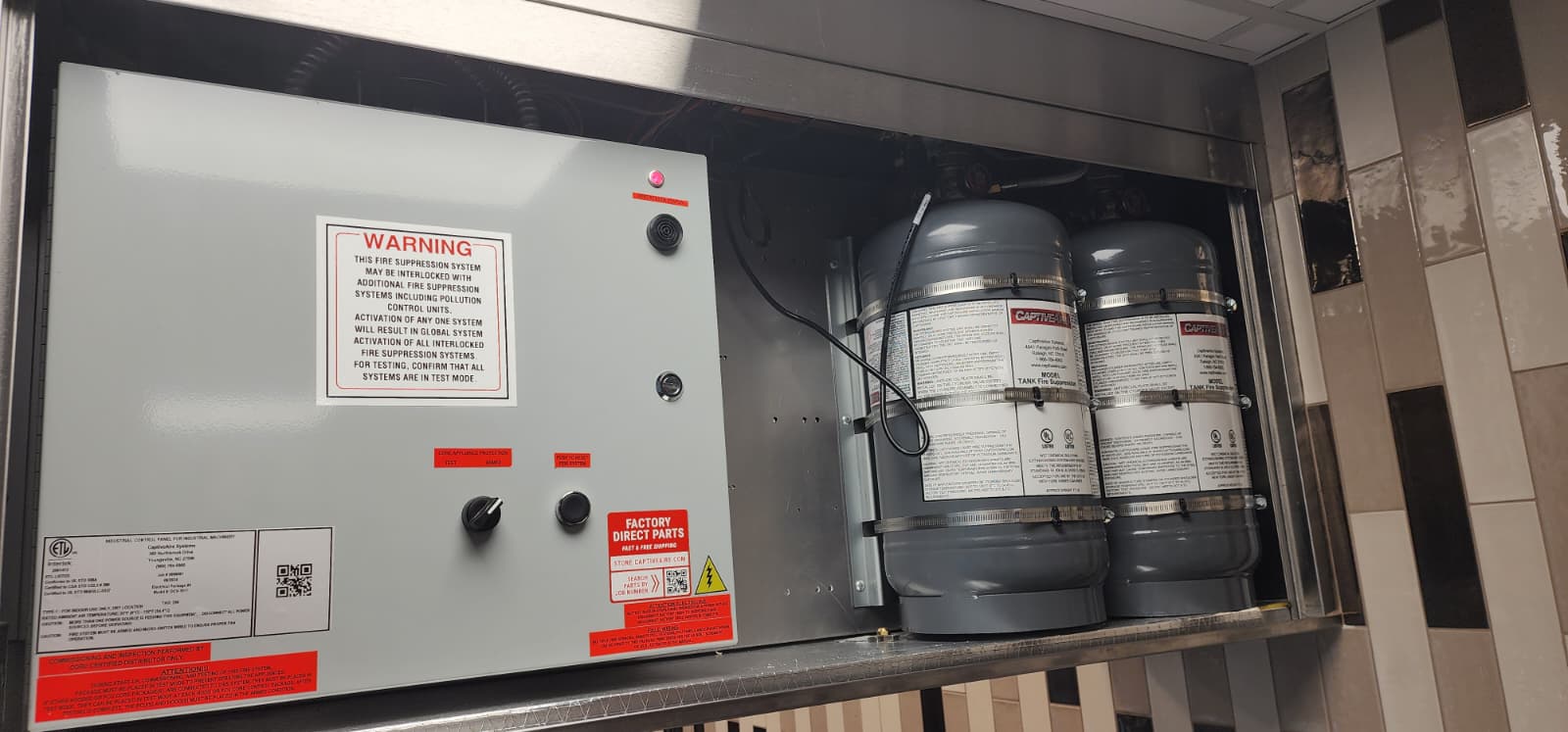 CaptiveAire kitchen suppression cabinet open: ETL-listed industrial control panel on the left with WARNING placard about interlocked systems and panel test mode, and a stainless cylinder bay on the right holding two grey CaptiveAire TANK wet-chemical cylinders
