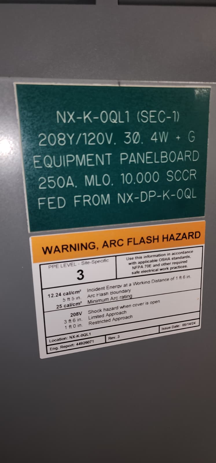 Kitchen branch electrical panel mounted on the wall above a kitchen prep area — grey panel cover with breaker handles inside and yellow/red label signage on the door identifying it as the panel feeding kitchen appliances under the hood, with breaker positions tied to the suppression-system shunt-trip outputs