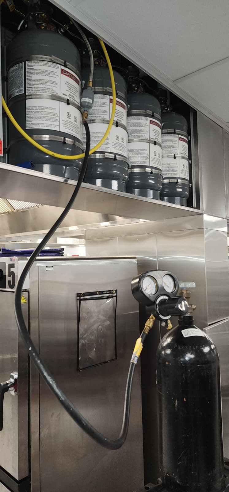 A CaptiveAire cylinder bay showing four agent cylinders mounted in line with their actuators connected by braided stainless pneumatic hoses, plus a tall nitrogen test cylinder standing alongside connected by a yellow regulated test hose