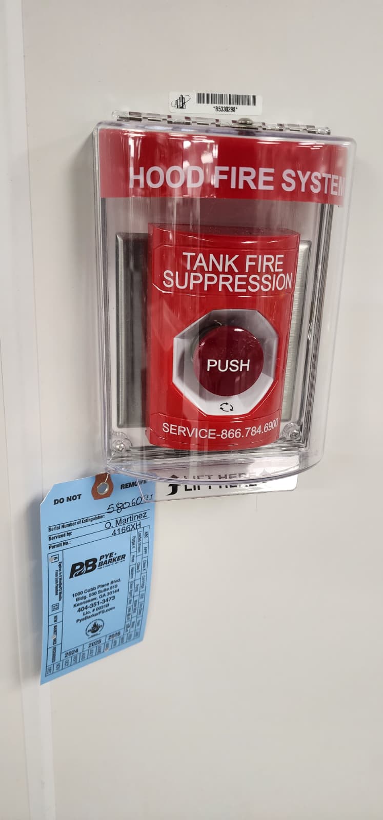 CaptiveAire manual pull station mounted on a kitchen wall — clear plastic protective cover labeled HOOD FIRE SYSTEM at the top, lifting open to expose the red TANK FIRE SUPPRESSION push-button with PUSH printed on the actuator and the SERVICE 866-784-6900 phone number underneath. A blue Pye-Barker semi-annual inspection tag hangs from the bottom edge with the service date, technician initials O. Martinez, and 2024 / 2025 / 2026 cycle punches
