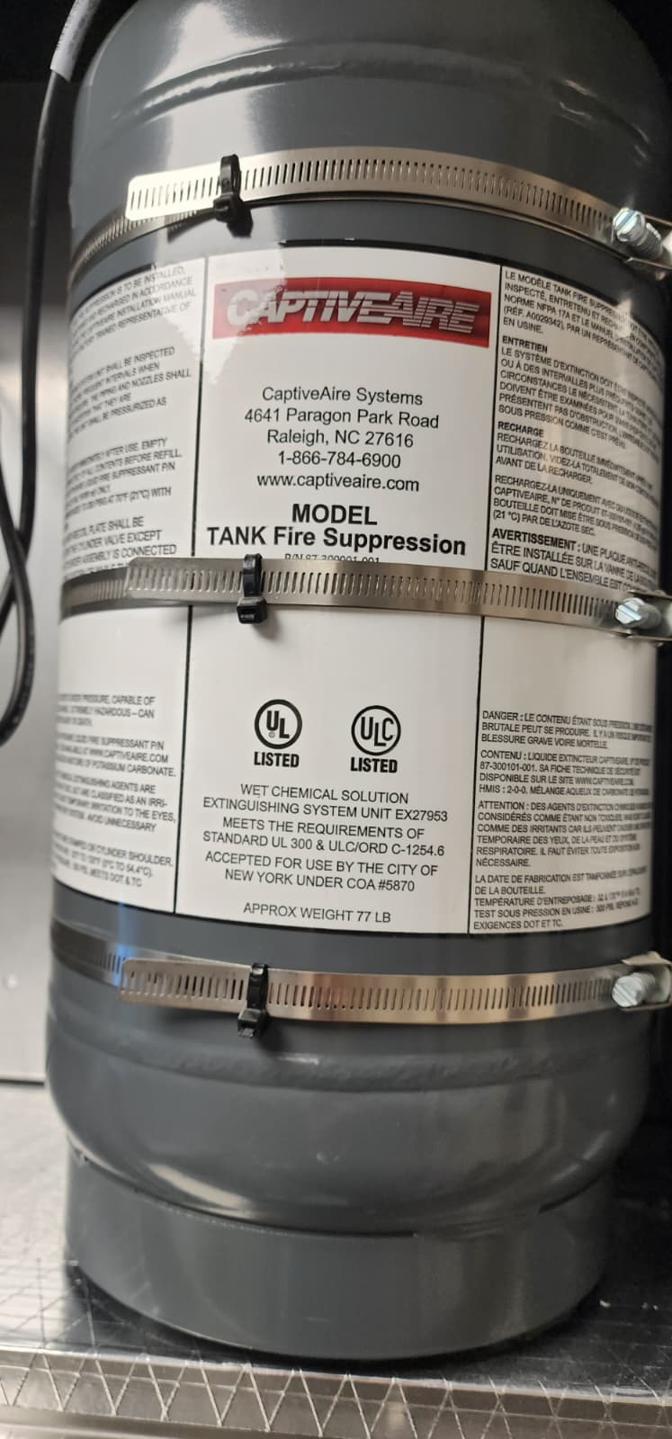 Close-up of a CaptiveAire TANK Fire Suppression cylinder label showing manufacturer address (Raleigh NC), 1-866-784-6900 service number, captiveaire.com, MODEL TANK Fire Suppression, UL Listed and ULC Listed marks, WET CHEMICAL SOLUTION EXTINGUISHING SYSTEM UNIT EX27953 meeting UL 300 and ULC/ORD C-1254.6, accepted for use by City of New York under COA #5870, approximate weight 77 LB, mounted on the cylinder body with three stainless steel bands
