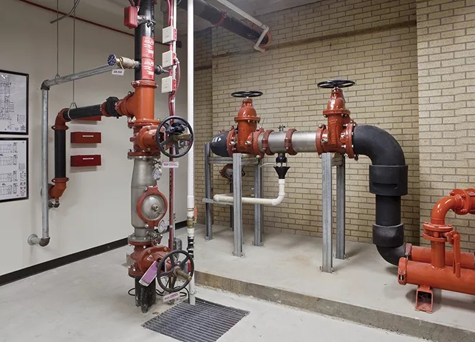 A fire protection backflow preventer room showing a reduced-pressure zone (RPZ) assembly on the left with its two OS&Y shutoff valves, test cocks, and a floor drain below the relief valve — alongside the main fire service header with grooved couplings feeding the building riser on the right