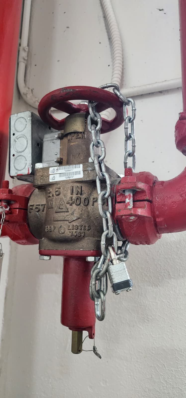 4½-inch 400 PSI brass-bodied control valve (butterfly pattern) on a red grooved-coupling fire protection riser — fitted with an electric tamper switch in a small junction box on the left, a red handwheel with 'OPEN' cast into the flange, and a chain-and-padlock keeping the valve mechanically locked in the open position
