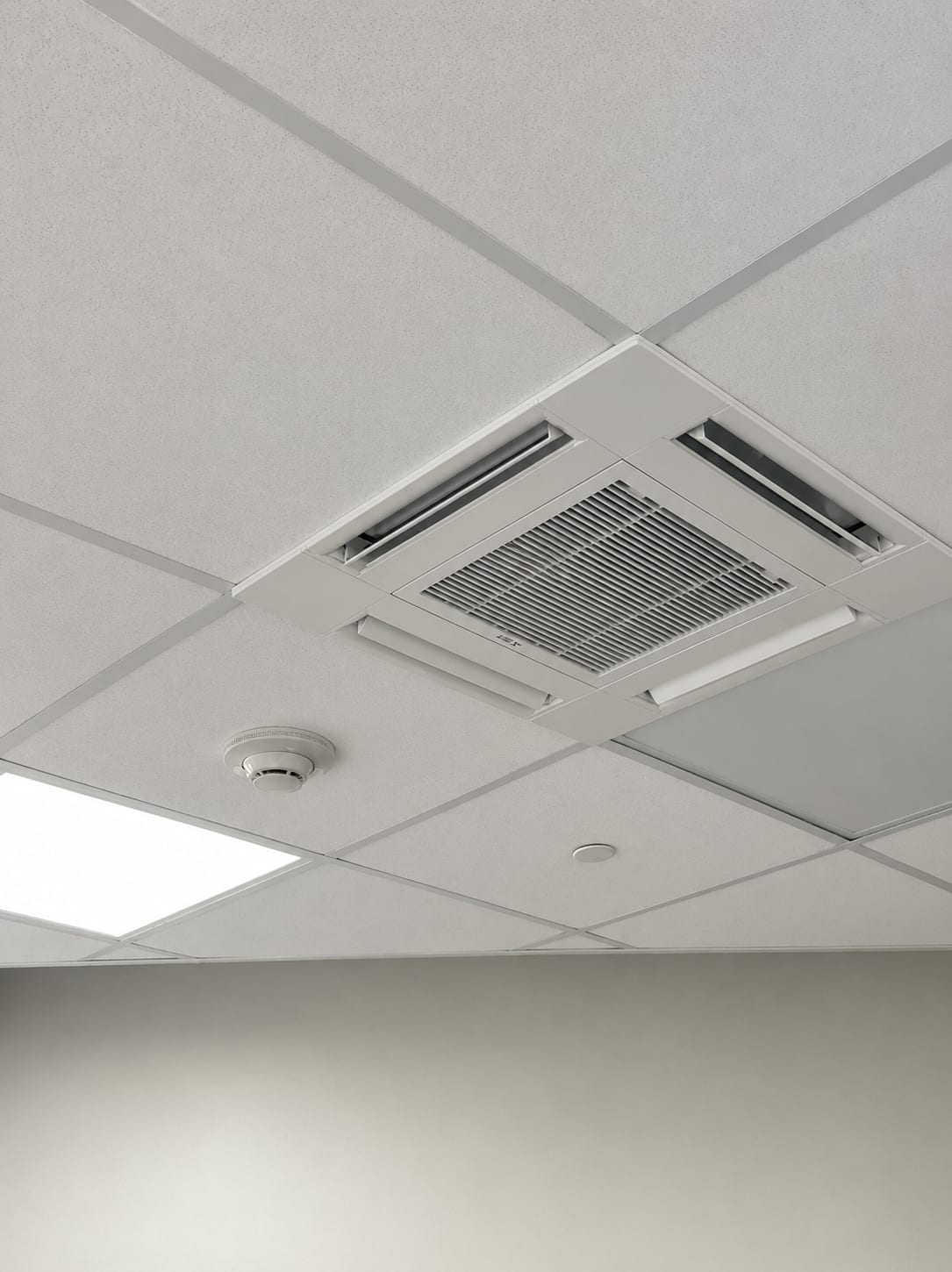 Ceiling-mounted four-way cassette HVAC unit with slot diffusers on each side and a center return grille, with a spot-type smoke detector installed on the directly adjacent ceiling tile