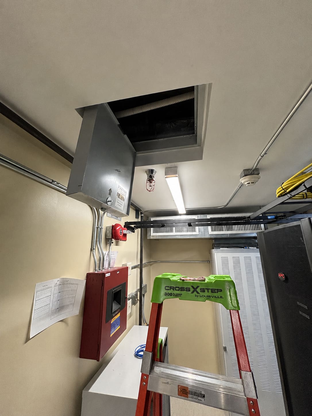 Ceiling-mounted fire-rated access hatch standing open above an equipment room, with a fire alarm control panel, alarm bell, sprinkler head, and a step ladder visible below the open hatch