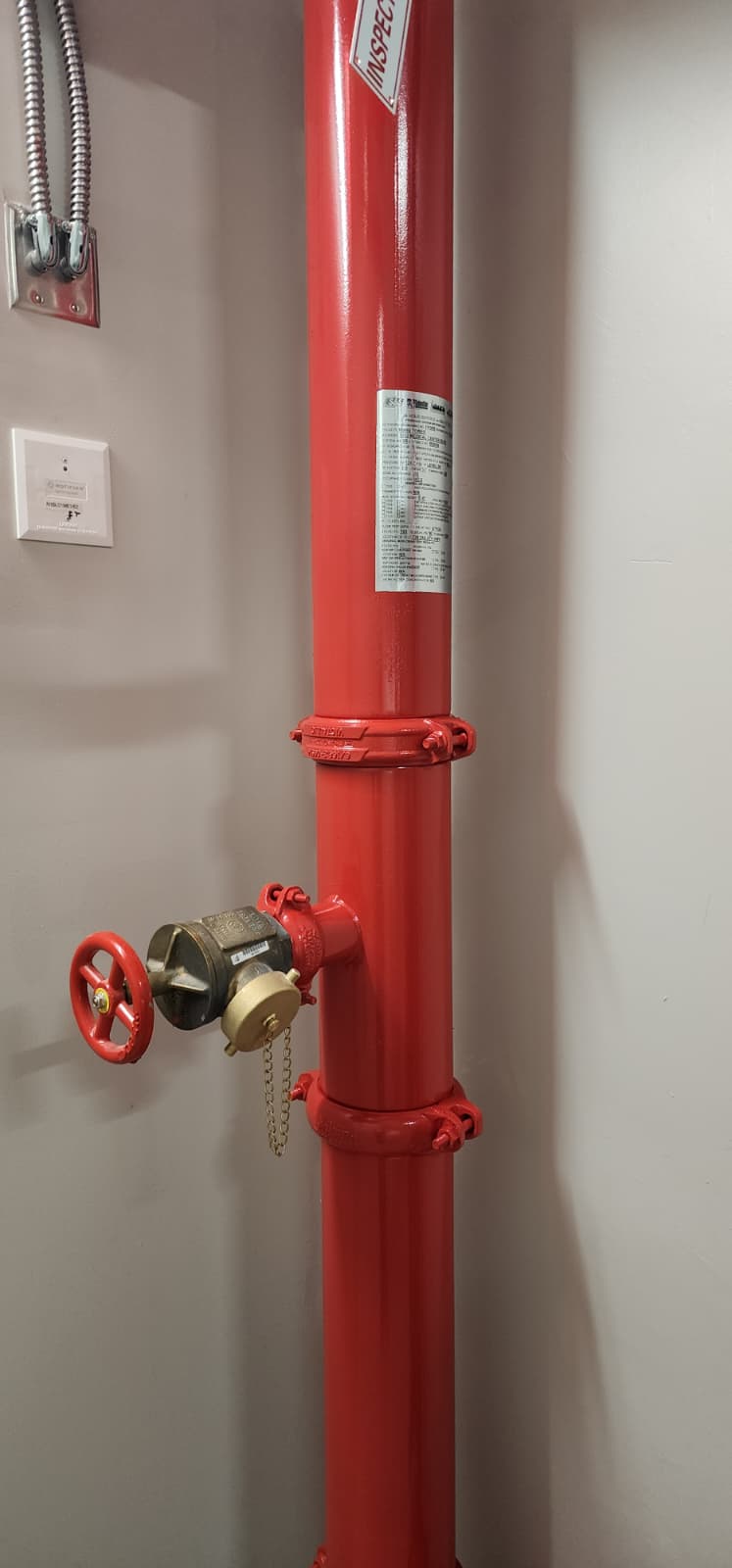 2½-inch Class I fire hose valve installed on a red-painted standpipe riser with brass valve body, chained cap, red handwheel, grooved couplings, and the standpipe identification label — located in a typical building stair/standpipe enclosure