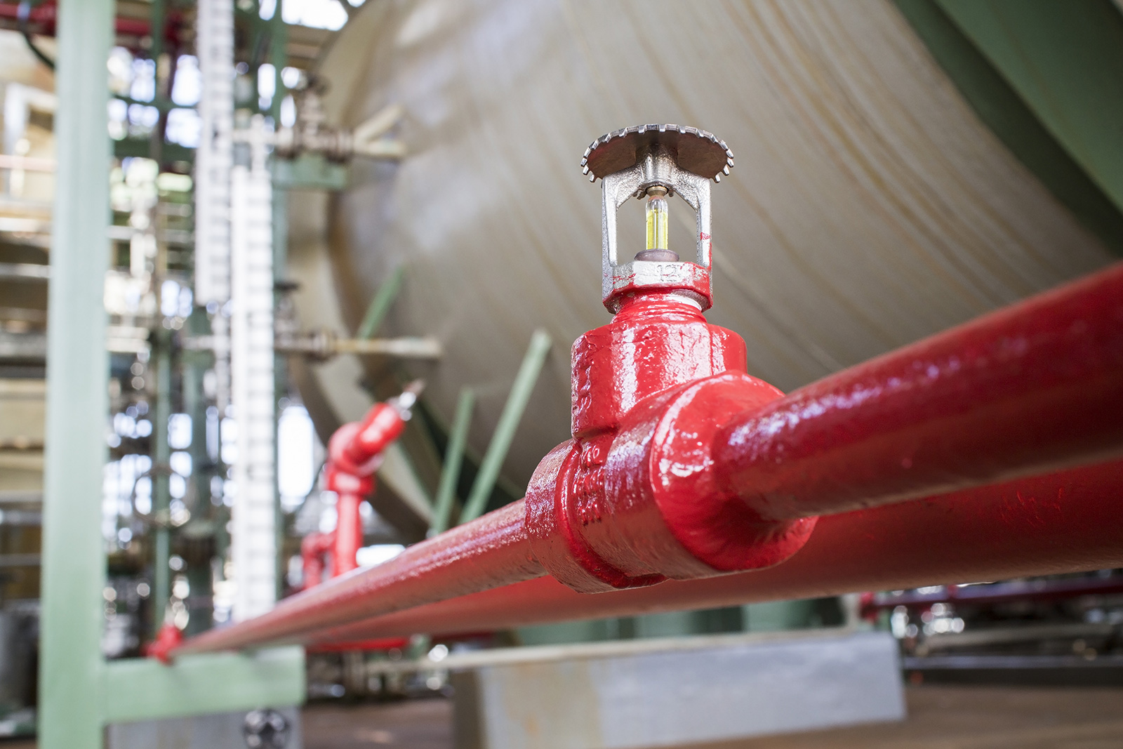 Close-up of an upright pendant-style fire sprinkler with a 212°F yellow glass bulb mounted on a red-painted branch line via a grooved tee fitting — industrial facility with storage tanks and additional sprinkler piping visible in the background