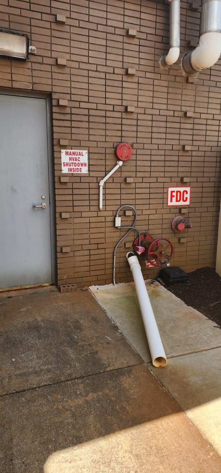 Field photo of a main drain valve and discharge on a fire sprinkler riser — the drain pipe exits to an exterior turned-down elbow