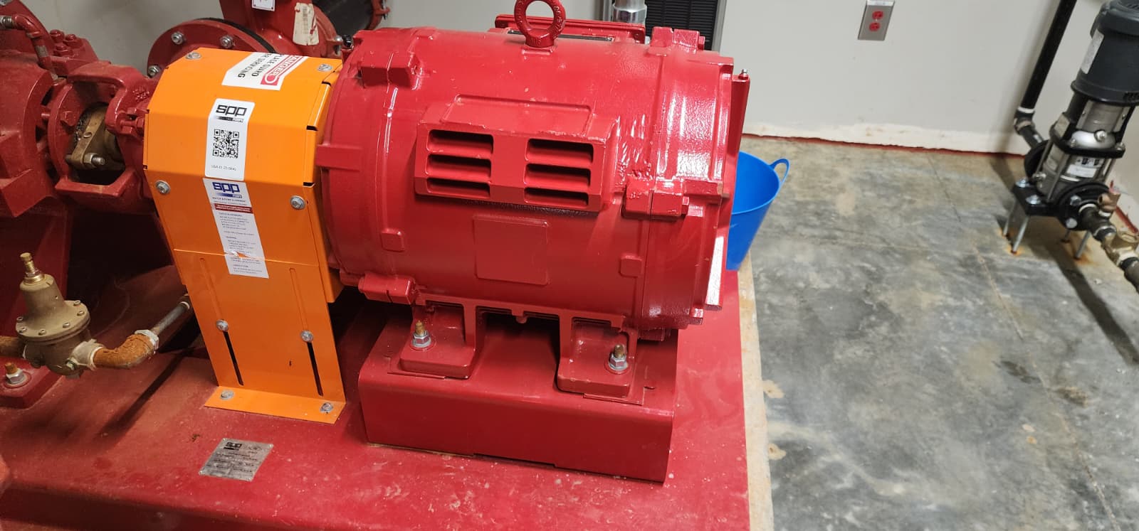 Red SPP fire pump electric motor close-up — TEFC housing with cooling vents, orange coupling guard with manufacturer QR code, vibration-isolation grouted baseplate, and flex conduit for motor feeder