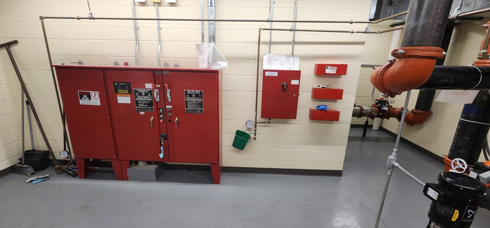 Complete fire pump room view showing electric fire pump controller, fire pump power transfer switch, jockey pump controller, spare sprinkler cabinets, gauges, a test header bucket, grooved discharge piping, and the electric pump motor