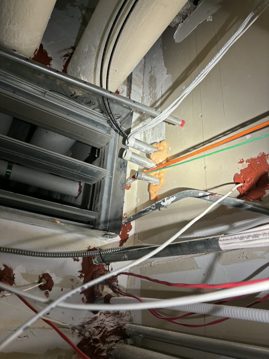 Orange fireblock foam blobbed around conduit penetrations in a fire-rated wall above the ceiling, next to properly installed red firestop sealant