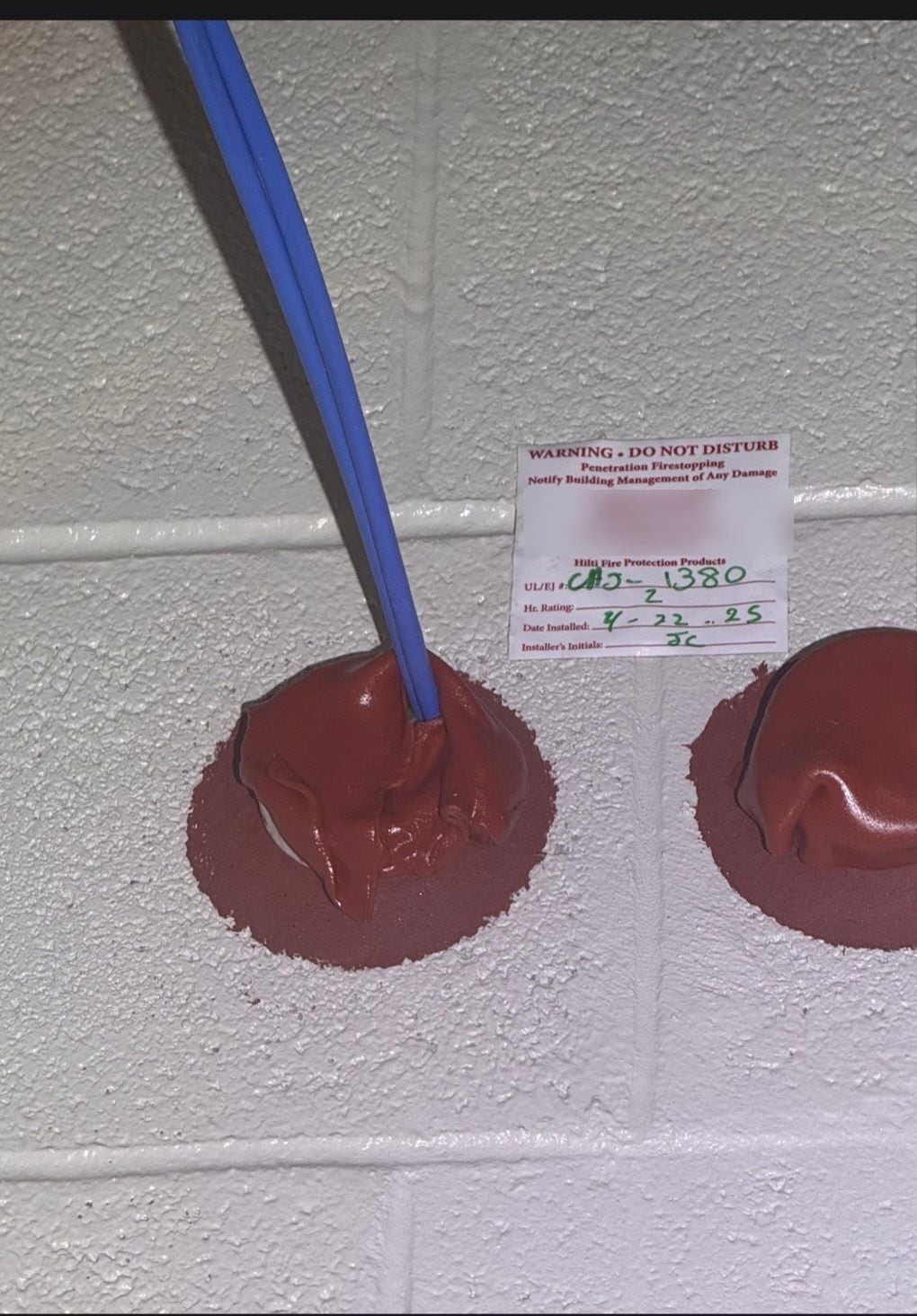 Two core-drilled penetrations in a CMU wall packed with red firestop putty, a blue jacketed cable passing through the left one, and a firestop label between them citing UL system CAJ-1380 with a 2-hour rating