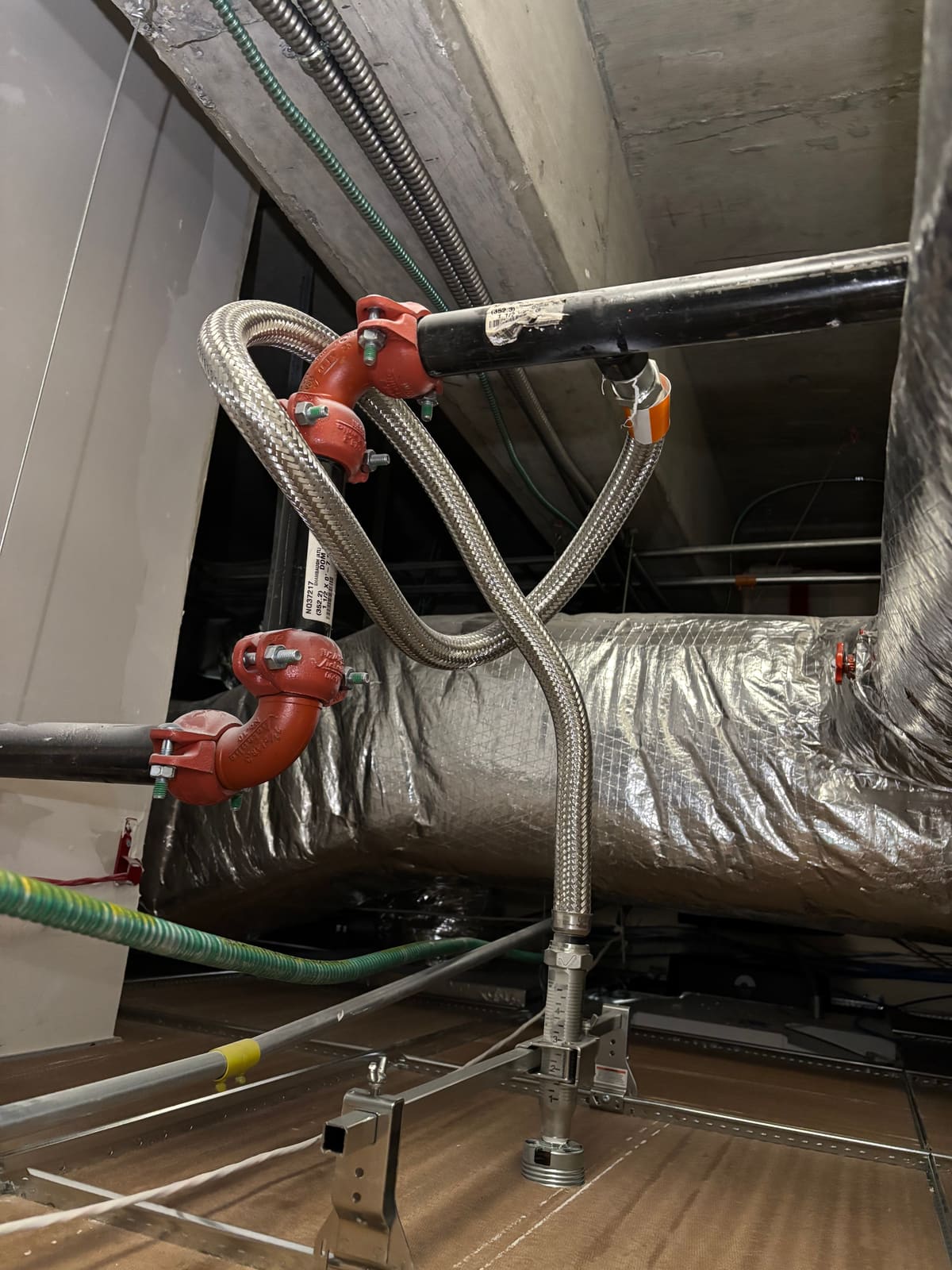Two braided flexible sprinkler hoses above a suspended ceiling — the left hose crosses over itself in a full loop threaded around the vertical drop pipe, the right hose sweeps over the ductwork in an S-curve