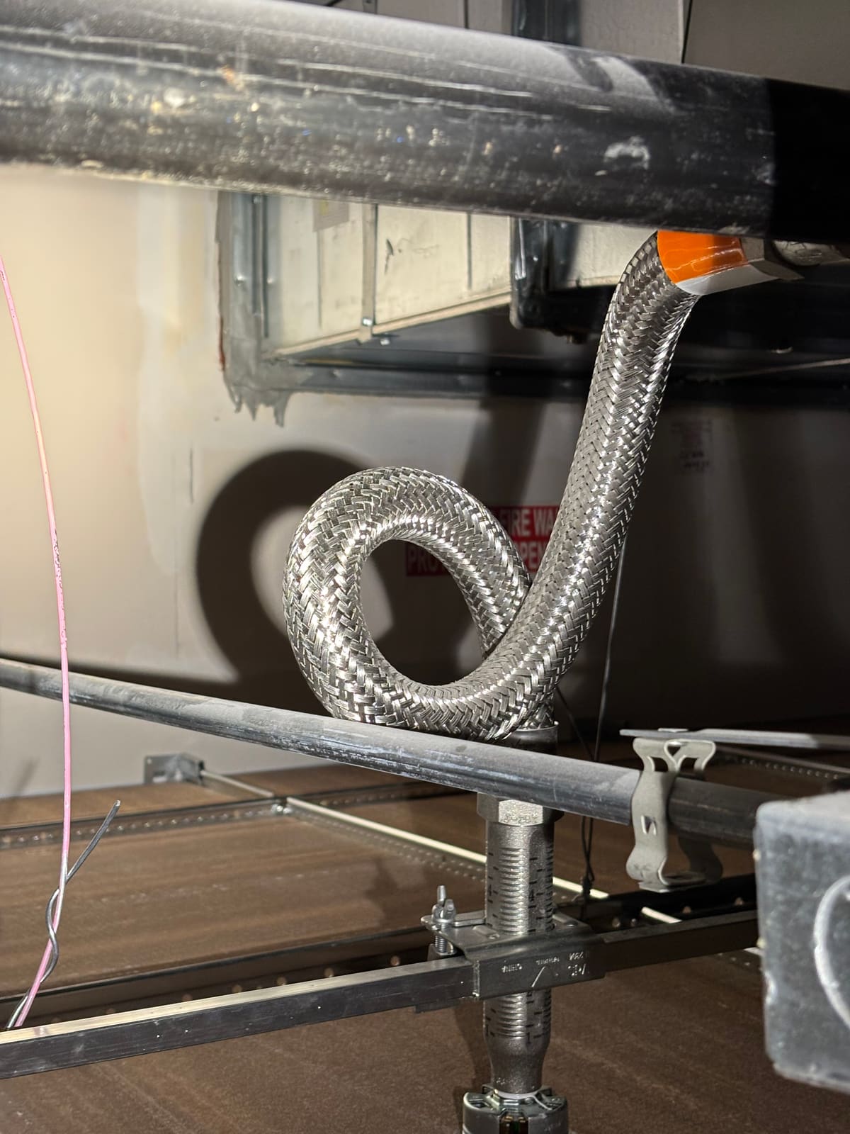 Braided flexible sprinkler hose curled into a tight pigtail loop directly above the drop, with the loop resting on a pipe below and a fire wall stencil visible on the wall behind