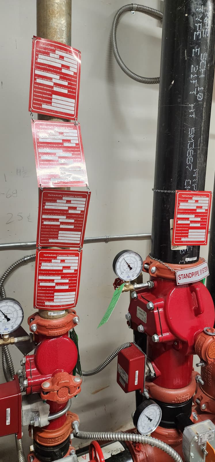 Multiple hydraulic design placards mounted on a sprinkler riser — four placards stacked vertically on one riser and additional placards on an adjacent standpipe system riser, with pressure gauges visible