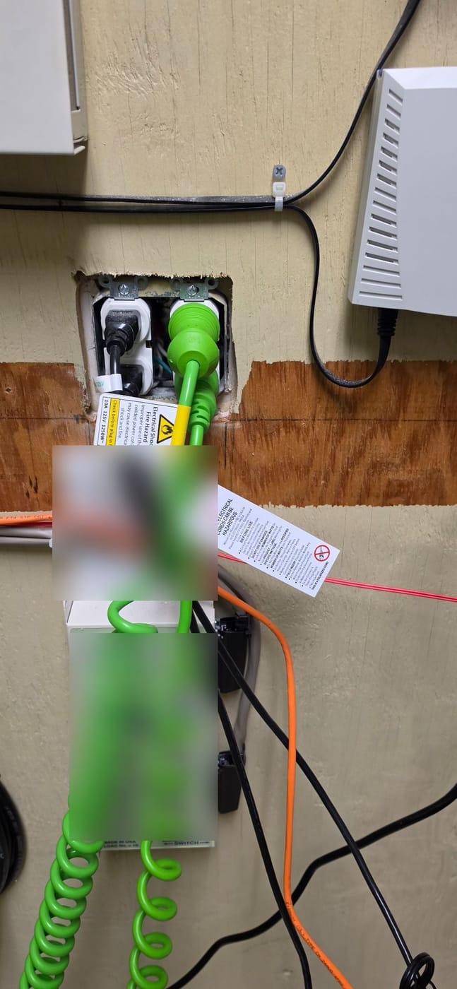 An open wall box with no cover plate and cords plugged into the exposed receptacles