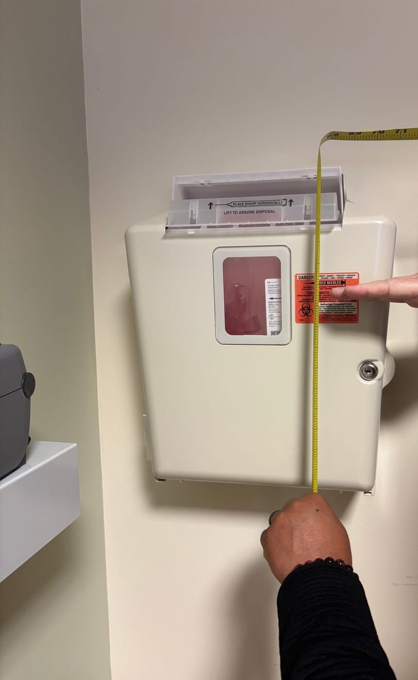 A tape measure held against a wall-mounted sharps container, showing how high its opening sits