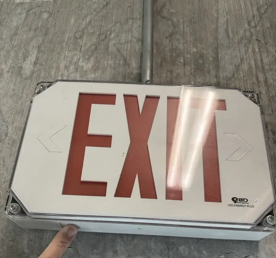Weatherproof LED exit sign mounted on a concrete wall in a parking deck stair, with a finger pressing the test button on the bottom edge of the housing
