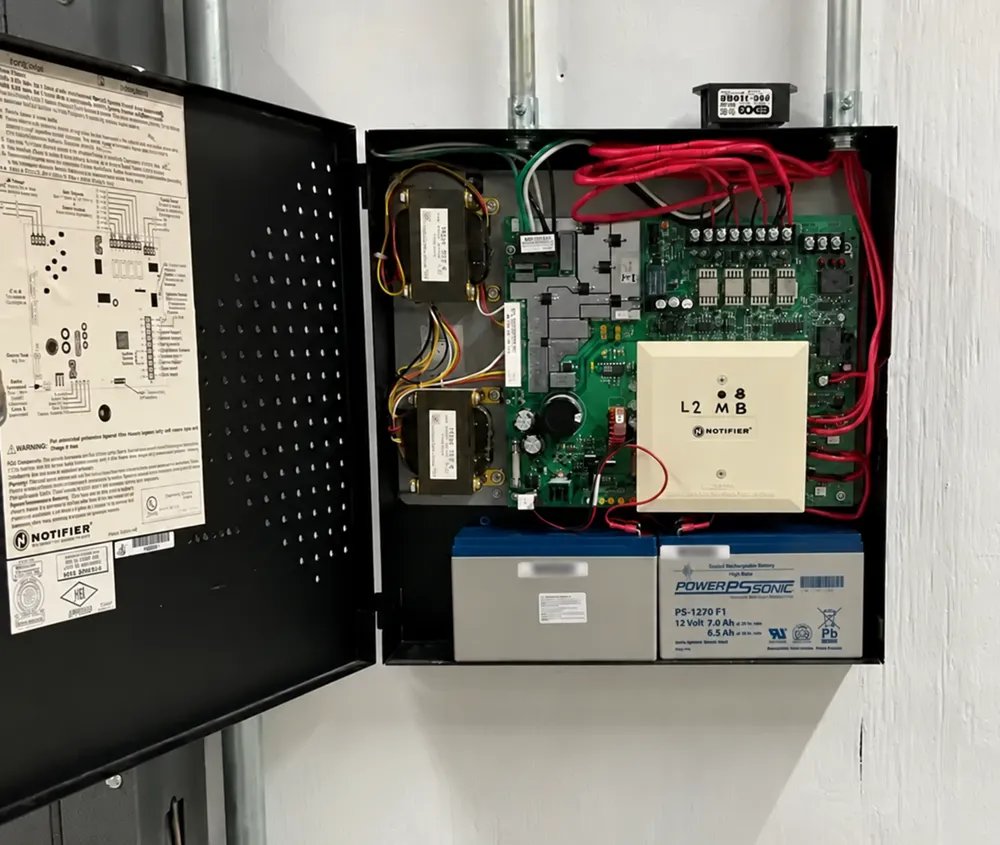 Open fire alarm power supply cabinet on a wall, with its circuit board, two transformers and an addressable module, and two 12-volt sealed lead-acid standby batteries of different makes sitting in the bottom of the cabinet