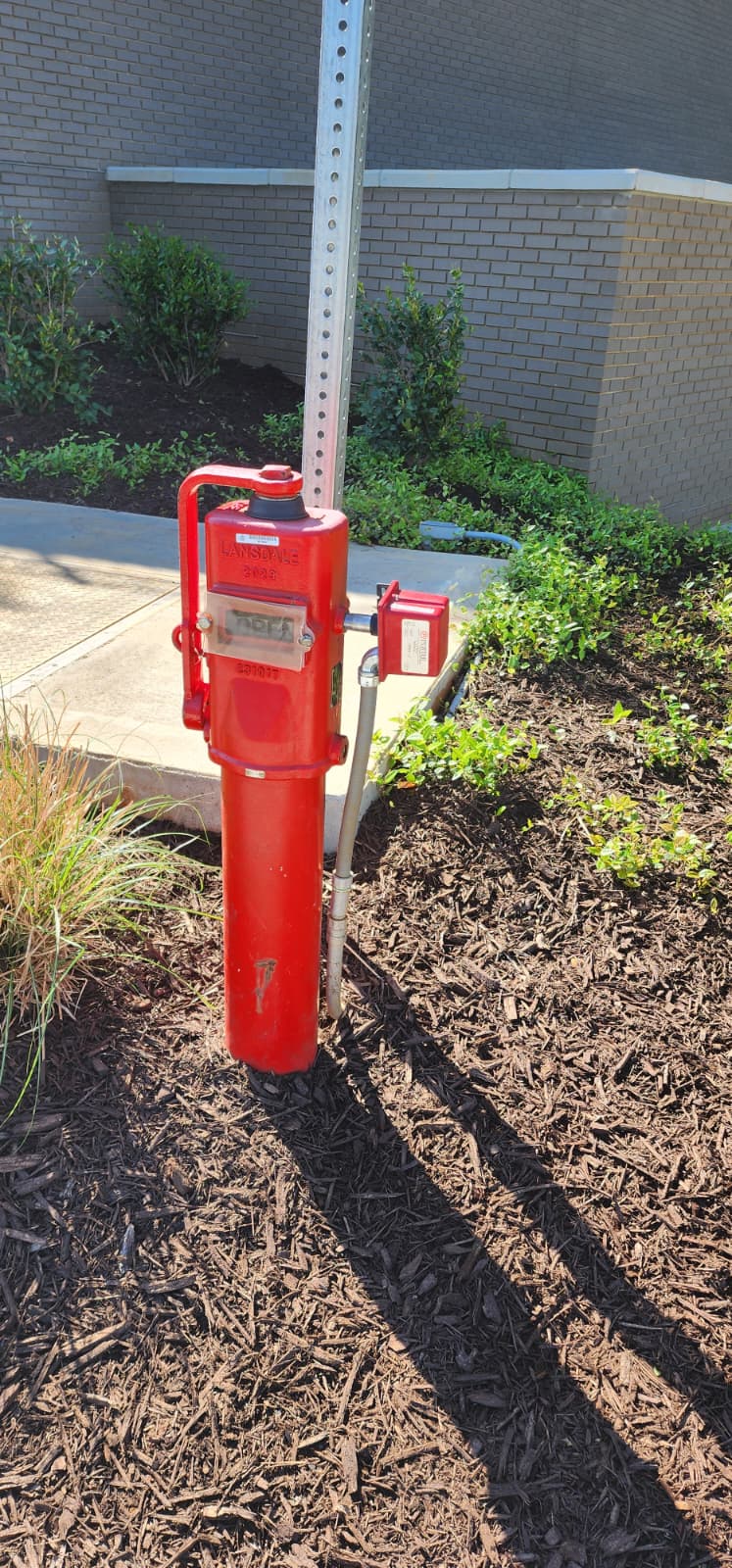 Close-up of a red post indicator valve with tamper switch housing and a padlocked operator nut, installed next to a metal signpost in a commercial landscape bed