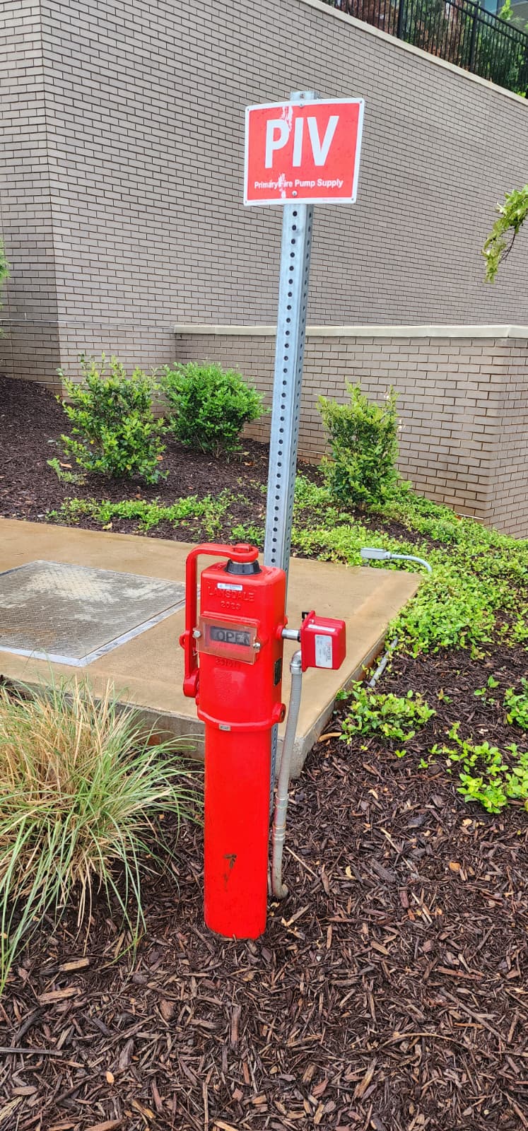 Classic freestanding post indicator valve — red Mueller post-style PIV with OPEN window indicator and adjacent tamper-switch housing in a landscaped bed, with a 'PIV — Primary Fire Pump Supply' identification sign on a metal post