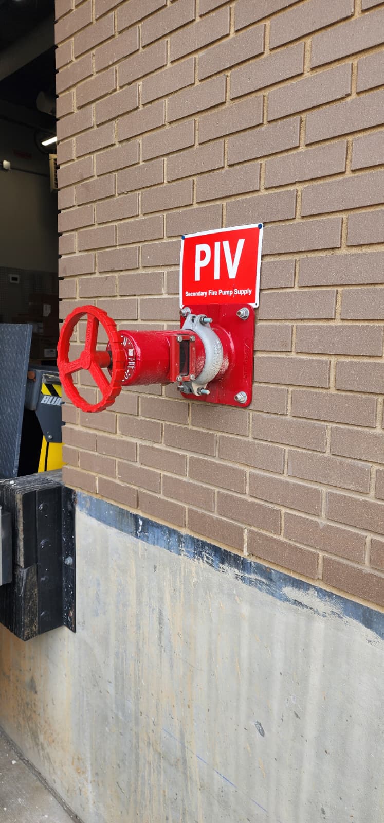 Wall-mounted Post Indicator Valve (WPIV) — red gate valve with handwheel mounted directly to a brick exterior wall, with rectangular window indicating valve position, with 'PIV — Secondary Fire Pump Supply' sign mounted above