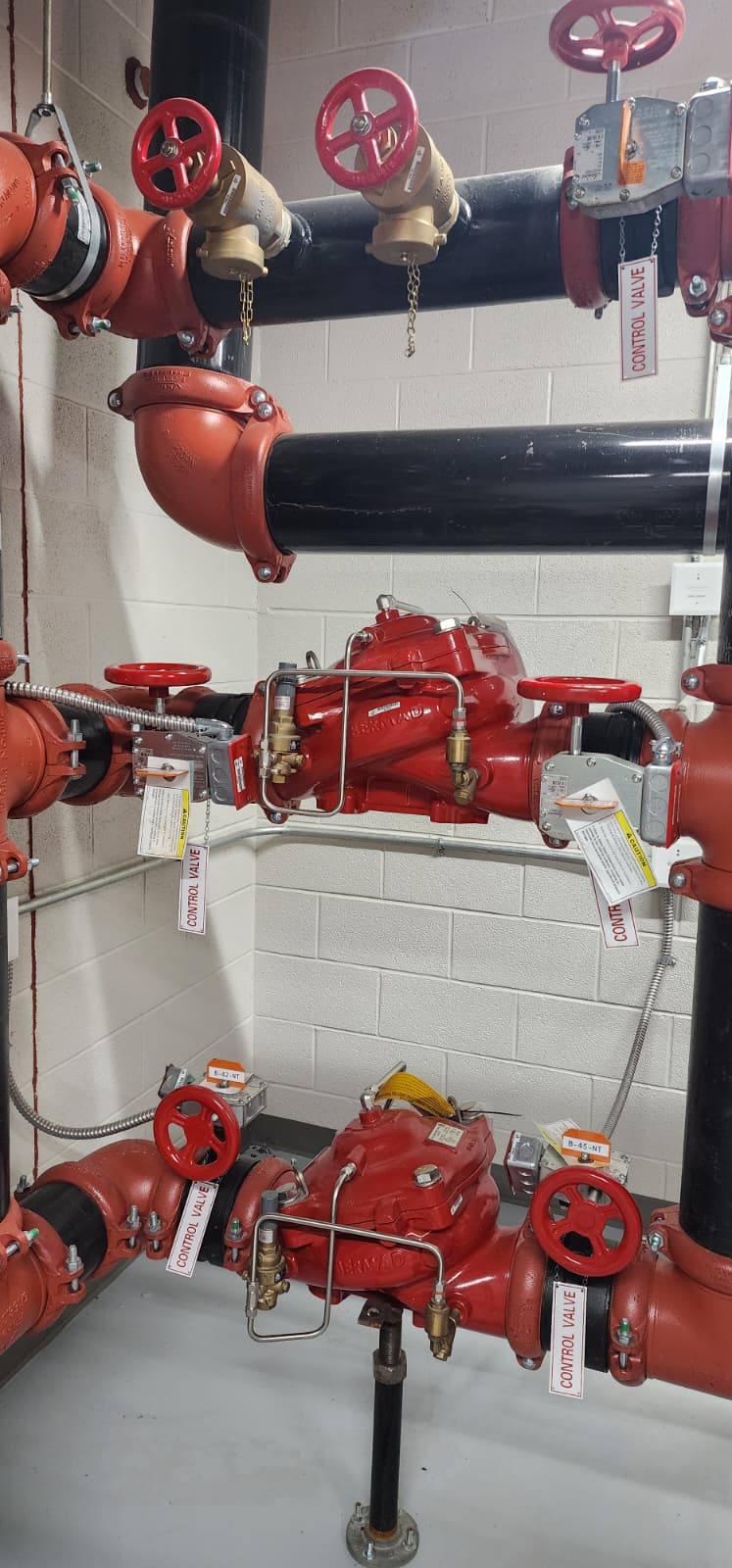 Two-tier stack of red Bermad pilot-operated pressure-reducing valves in a high-rise standpipe riser room, with sensing lines and isolation OS&Y control valves on either side, monitor modules with control-valve tags, and grooved couplings on the inlet and outlet pipes