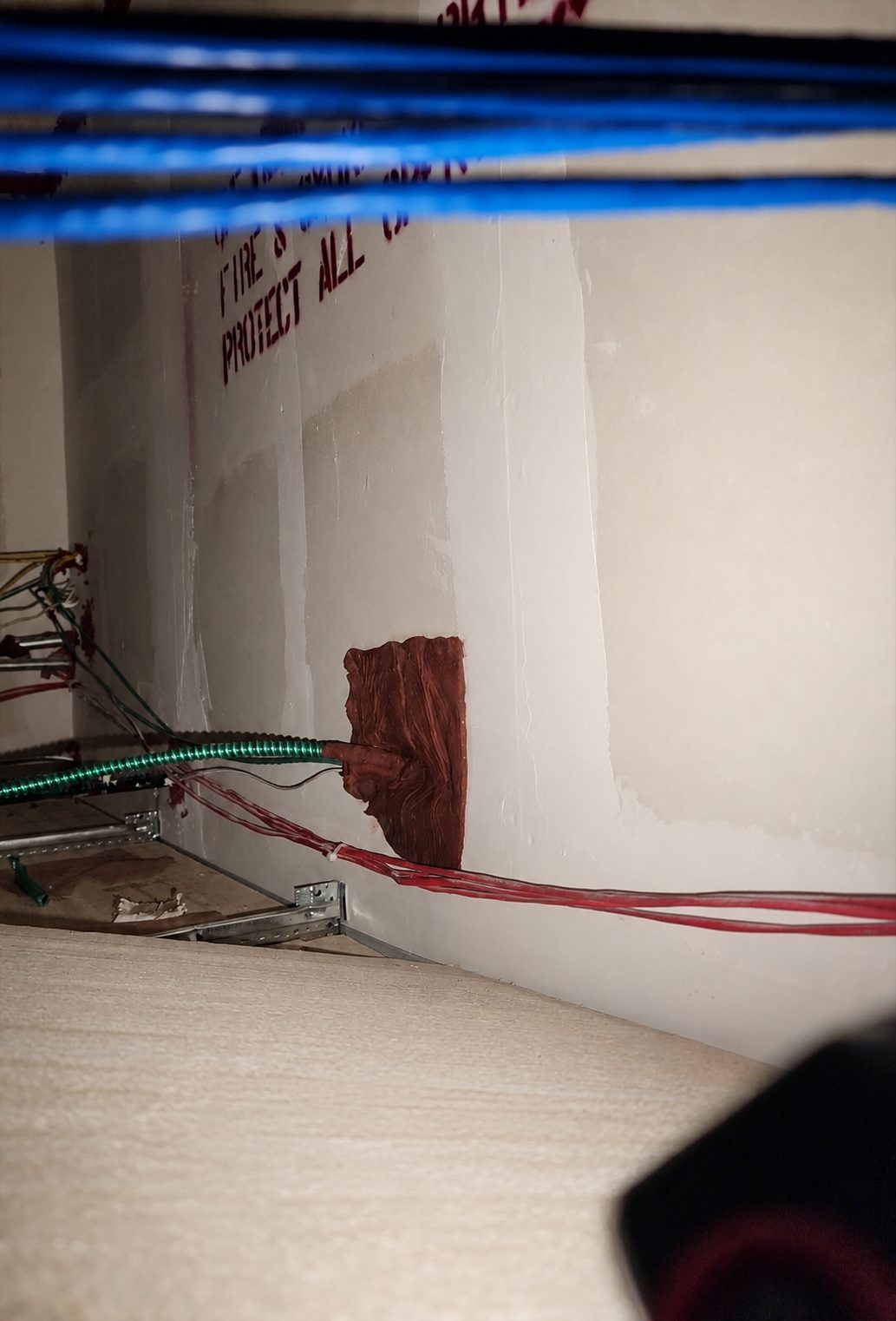 Fire-rated wall above ceiling with FIRE STOP PROTECT ALL marking visible — putty pad pressed over cable penetration with MC cable and wiring passing through
