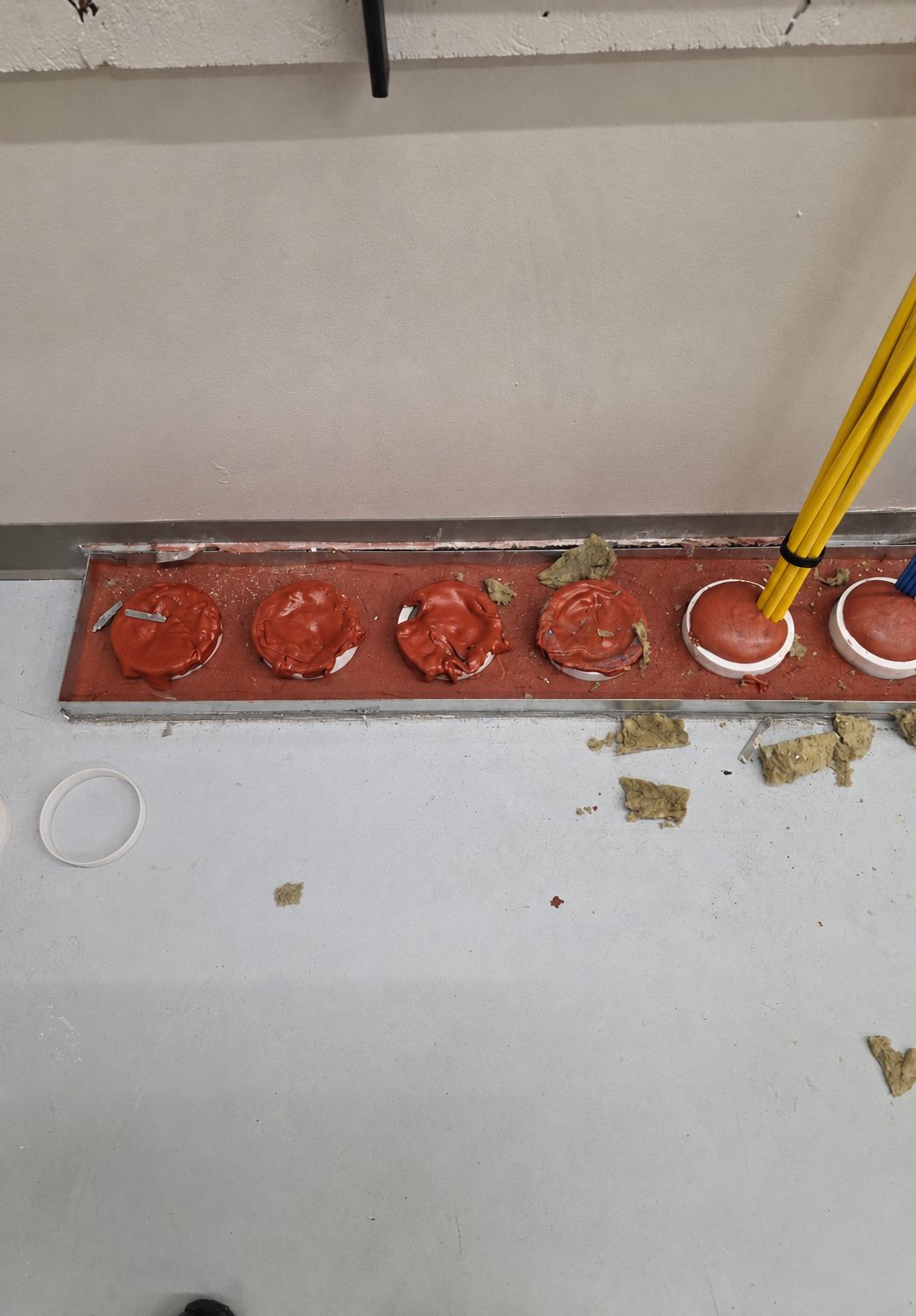 Floor penetration with multiple pipes passing through a metal sleeve — putty pad material blobbed over each pipe, mineral wool debris scattered around