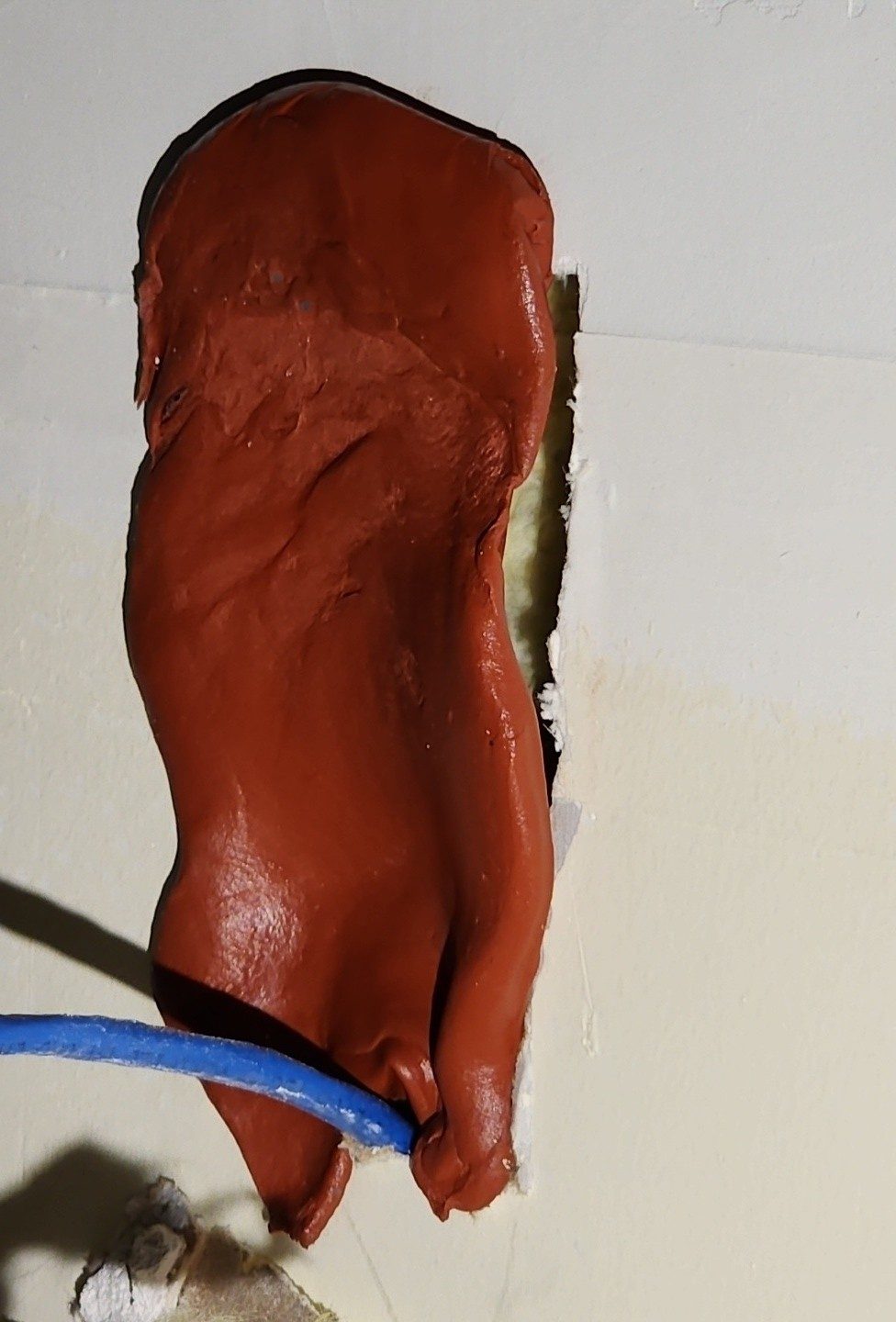 Putty pad material pressed over a wall penetration with a blue cable exiting the bottom — used as a drywall patch, not wrapping an electrical box