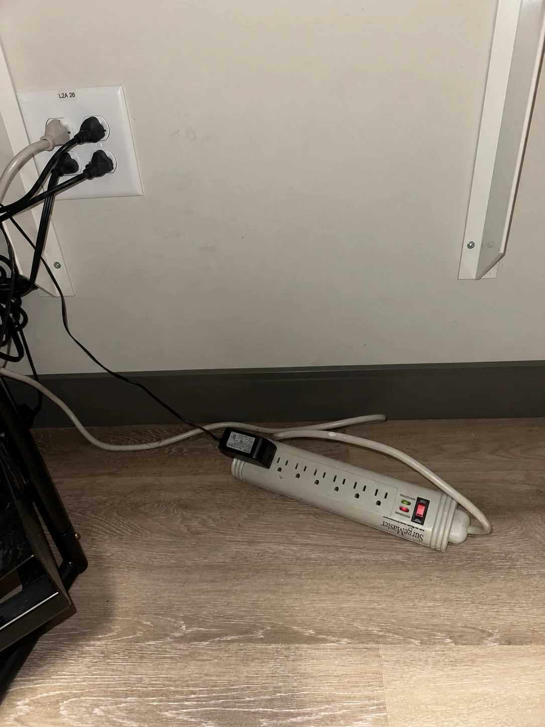A white SurgeMaster power strip lying flat on the floor, outlets facing up, its cord running loosely to a wall outlet that already carries a four-way adapter
