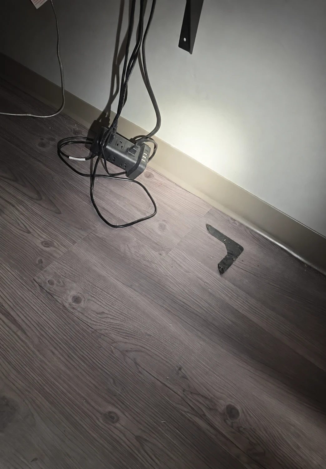 A tangle of black cords looping down to a power strip sitting on the floor against a baseboard, with a loose metal mounting bracket also lying on the floor nearby
