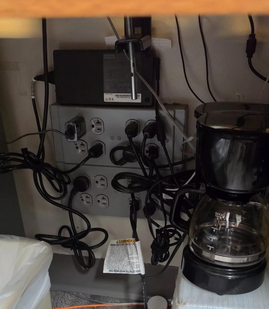 Two stacked gray multi-outlet modules mounted under a desk, a laptop power brick clamped above them, a coffee maker beside them, and a nest of cords plugged into every receptacle