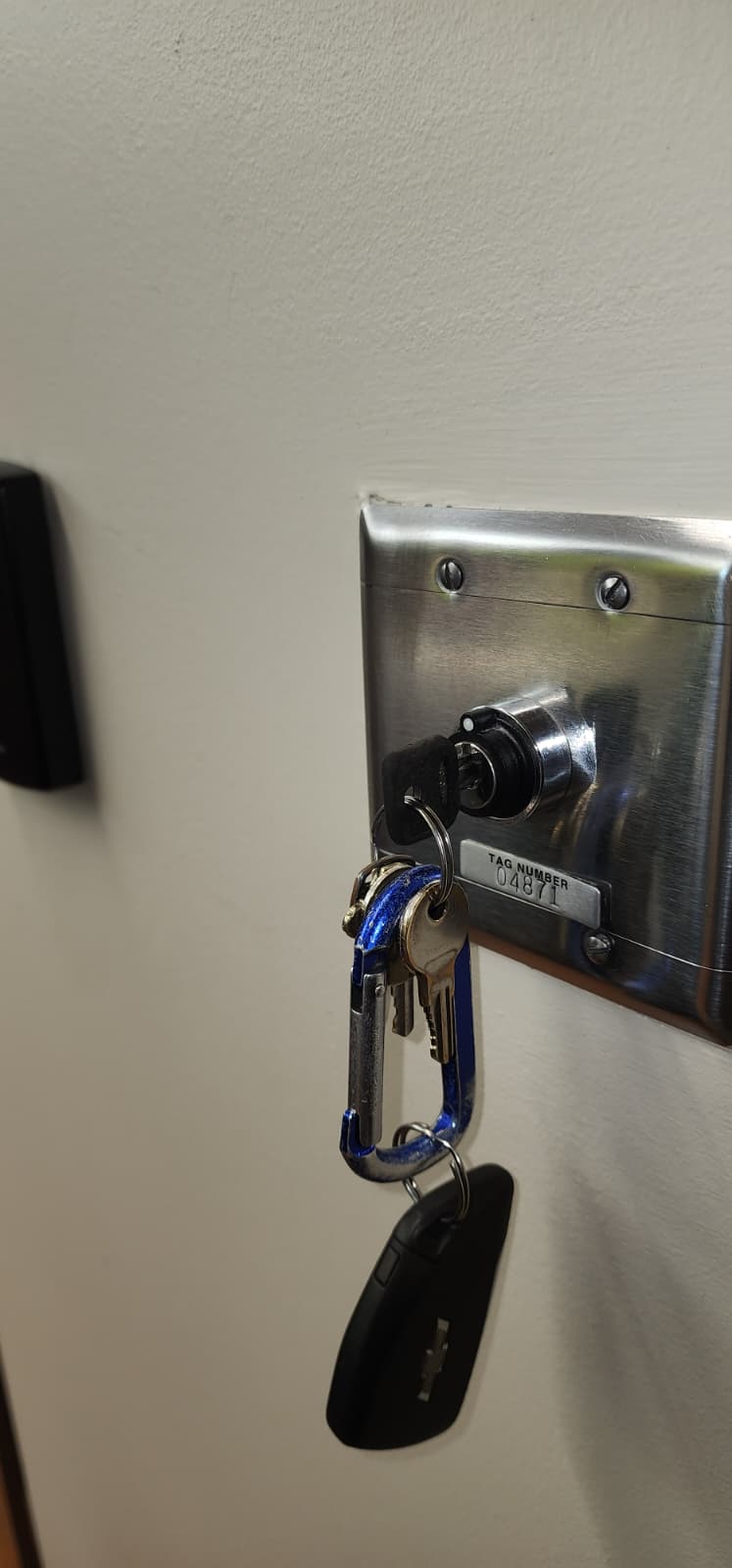 Stainless steel wall-mounted fire door key switch plate with a TAG NUMBER 04871 label and a key hanging in the switch cylinder