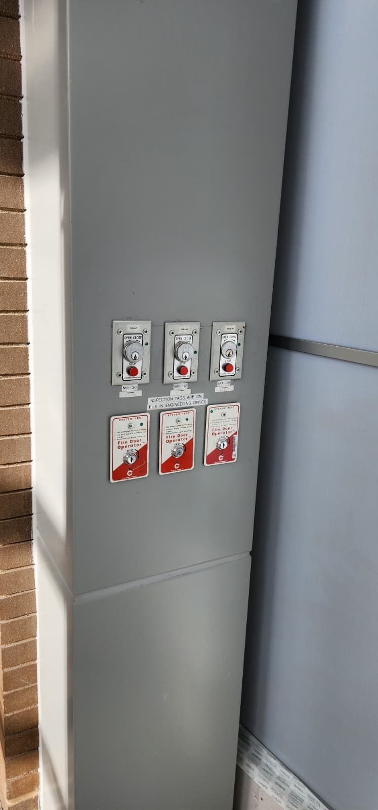 Column with three fire door key-switch wall stations labeled M1-36, M1-37, and M1-38, each with an annual Fire Door Operator TEST inspection tag below it, and a sign noting inspection records are on file in engineering