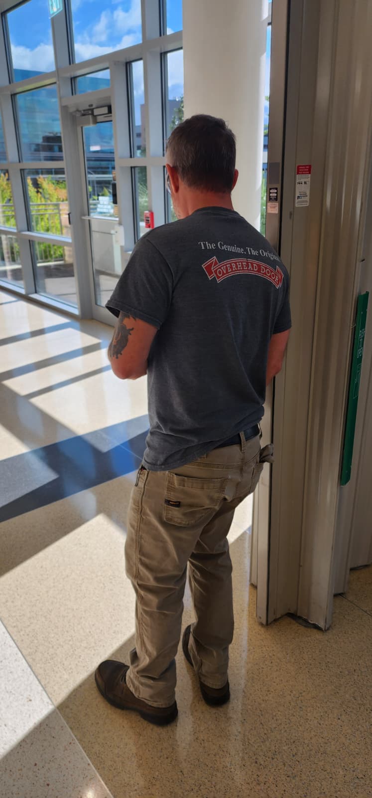 An Overhead Door technician performing an annual fire door inspection, standing at the leading edge of the door with the annual drop test record sticker visible on the door frame