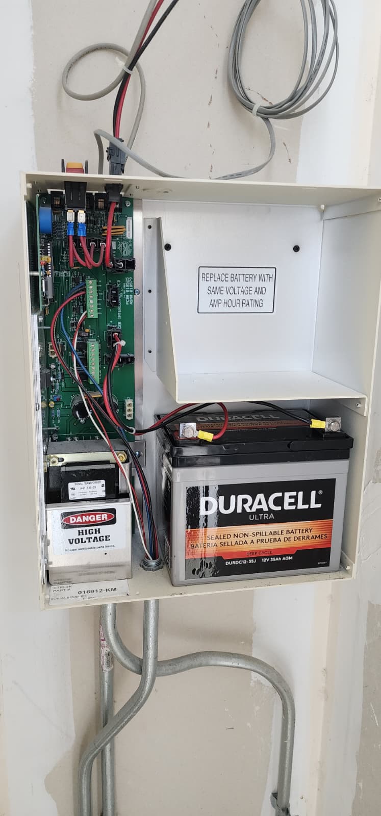 Interior of a WON-Door operator controller enclosure showing a Duracell 12V 35Ah AGM sealed non-spillable battery, transformer, and circuit board with a DANGER HIGH VOLTAGE warning label