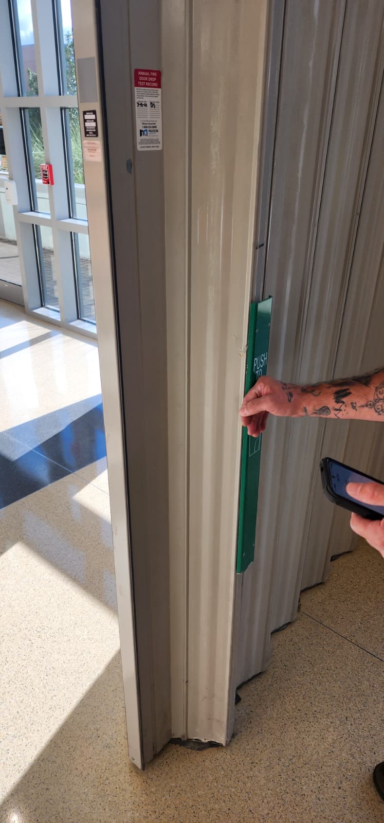 A person's hand pressing the green PUSH TO OPEN emergency egress bar on the leading edge of a WON-Door accordion fire door panel, demonstrating that the door can be manually pushed open from the egress side