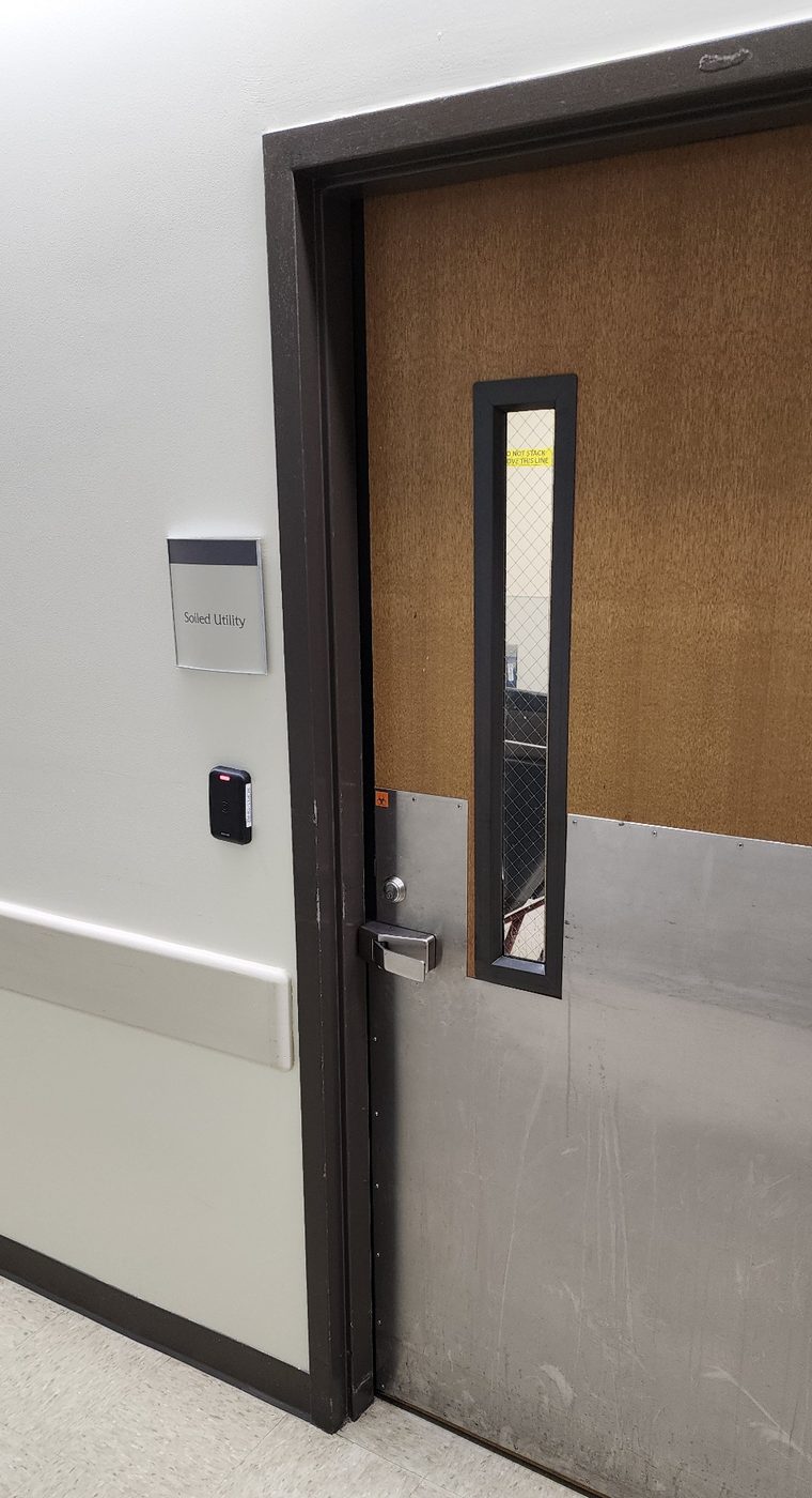 A closed soiled utility room door with a wired-glass vision panel, a stainless kick plate, a biohazard label near the latch, a wall sign reading Soiled Utility, and a badge reader beside the frame. A yellow sprinkler clearance sign reading DO NOT STACK ABOVE THIS LINE is visible through the window.