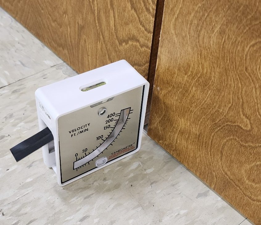 A Dwyer Vaneometer swing-vane anemometer sitting on the floor at the undercut of a closed soiled utility room door. Its scale is marked in feet per minute from 0 to 400 with a spirit level bubble on top, and the vane is deflected by air moving out of the room into the corridor — the wrong direction for a soiled room.