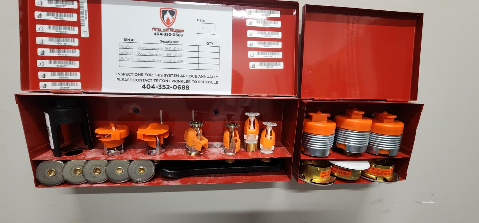 Triton Fire Solutions spare sprinkler cabinet opened showing inventory sheet, pendants, uprights, sidewalls, and concealed heads with orange protective caps and barcode tags