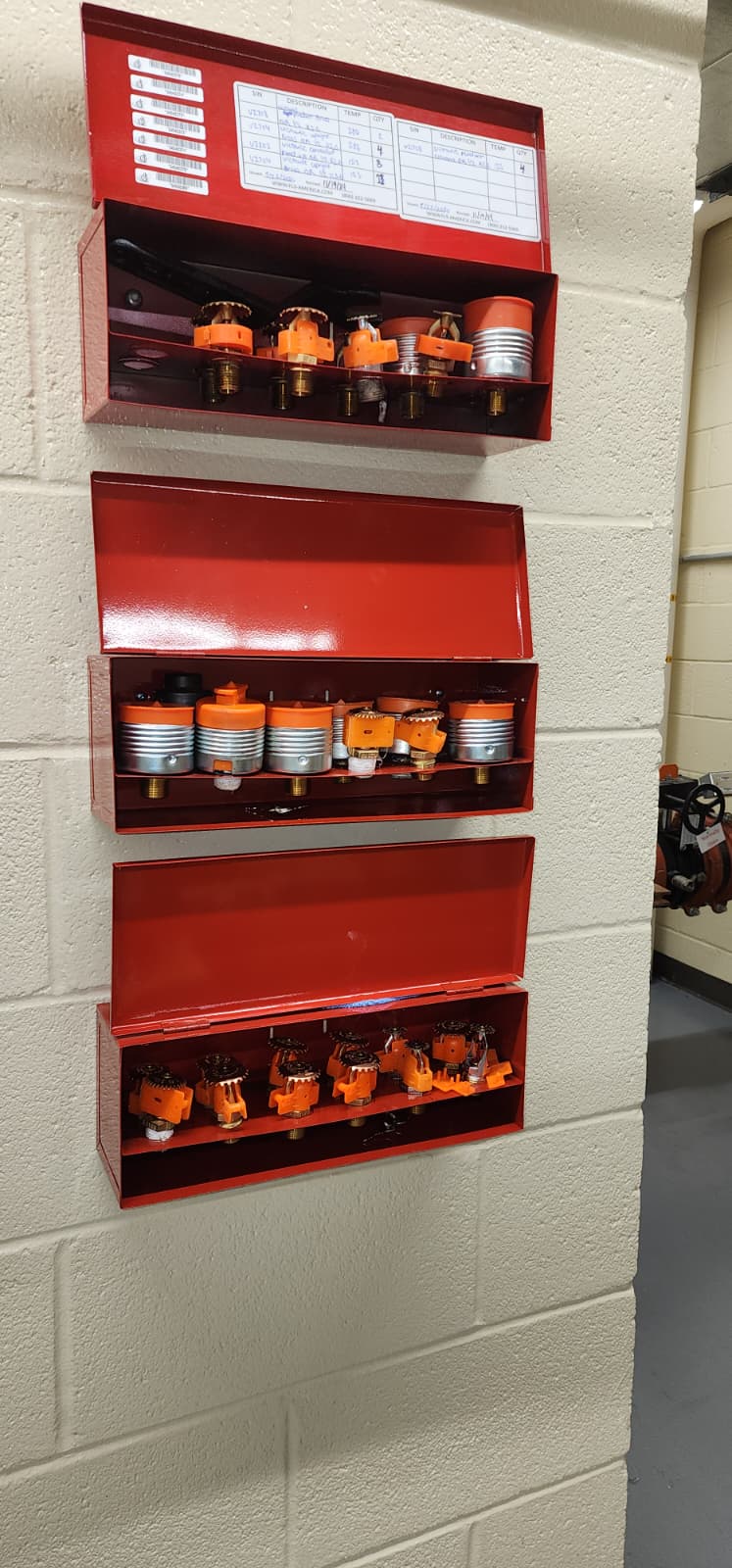 Three red spare sprinkler cabinets wall-mounted in a riser room — top cabinet open showing wrench, sprinkler inventory list, and multiple spare heads of different types and temperature ratings with orange protective caps