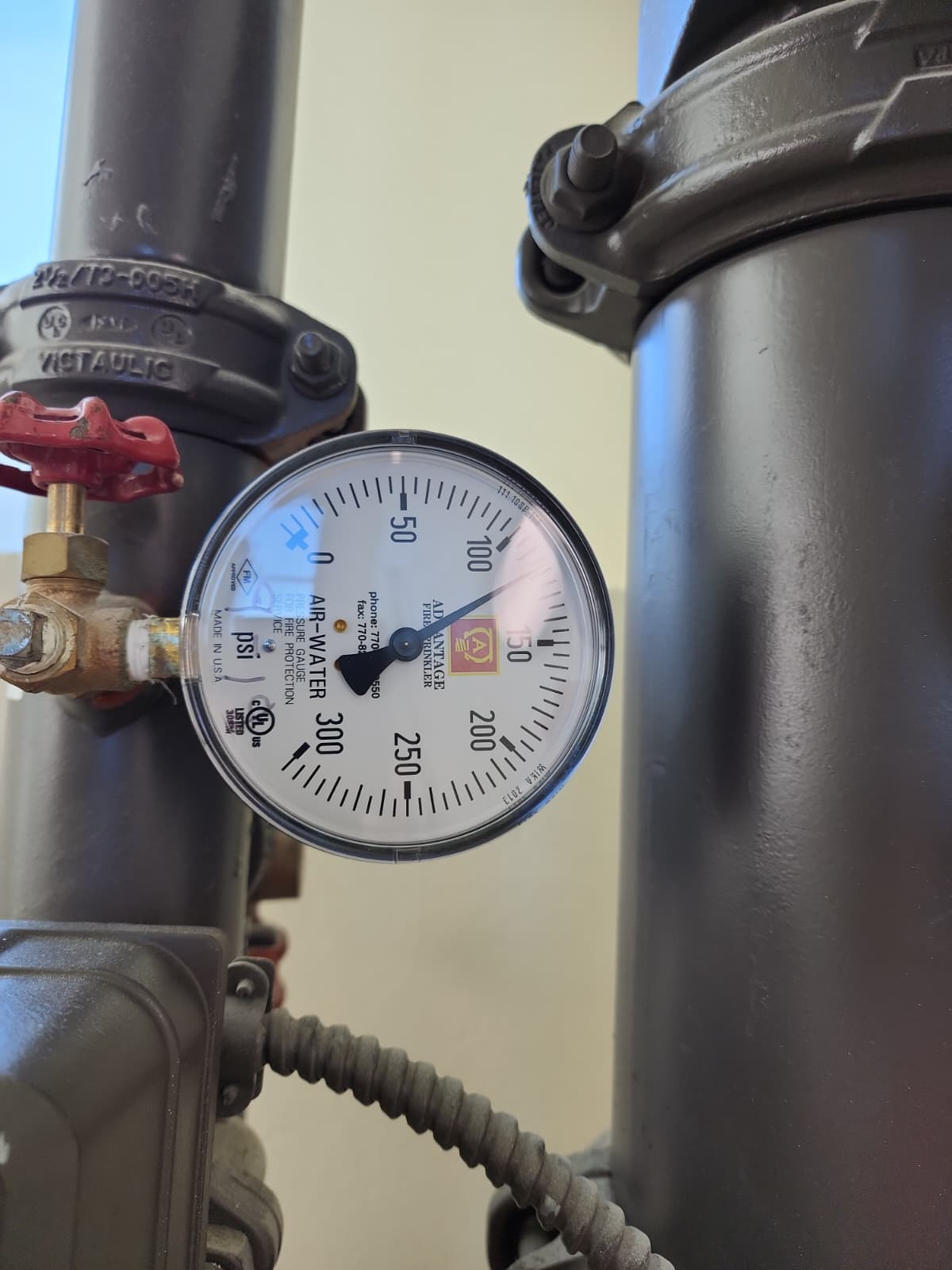 Advantage Fire Sprinkler 0–300 PSI dual-service AIR-WATER pressure gauge mounted on a sprinkler/standpipe riser assembly — brass needle-valve gauge isolation valve on the left with a red handwheel, Victaulic grooved couplings on the adjacent risers, flexible metallic conduit running down to a pressure switch or supervisory device below, FM-approved and UL-listed markings visible on the dial face
