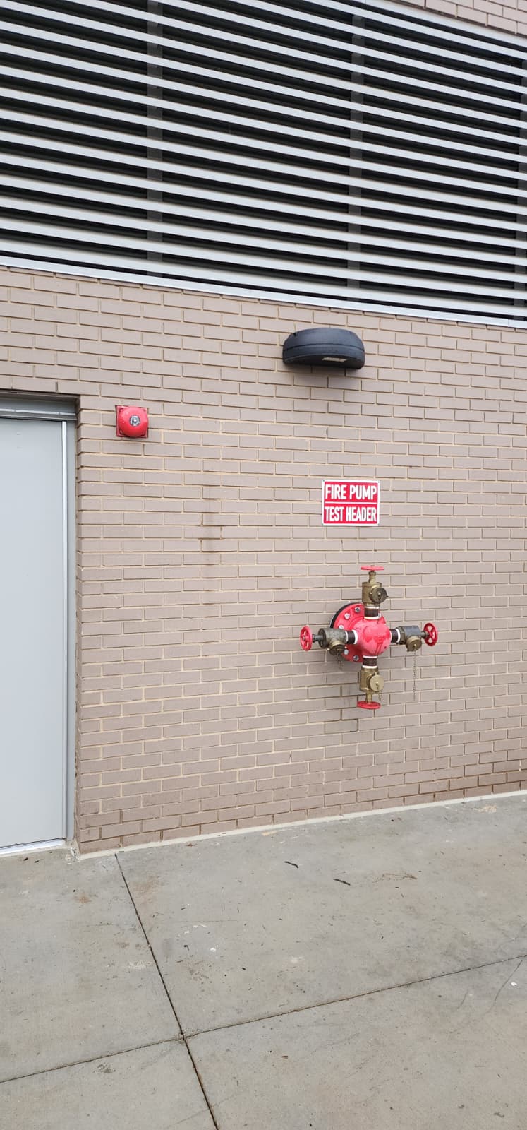 Real exterior building wall with a fire pump test header — three-outlet brass manifold with red handwheels and threaded discharge couplings, mounted to a brick wall below a labeled FIRE PUMP TEST HEADER sign and a supervisory bell, with a louvered ventilation grille above and a pump-room access door to the left