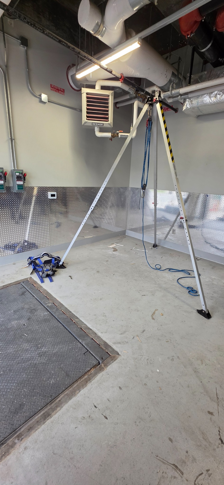 A portable aluminum tripod anchor rigged with a self-retracting lifeline over a floor access hatch, with a blue full-body harness staged on the floor beside it in a mechanical room