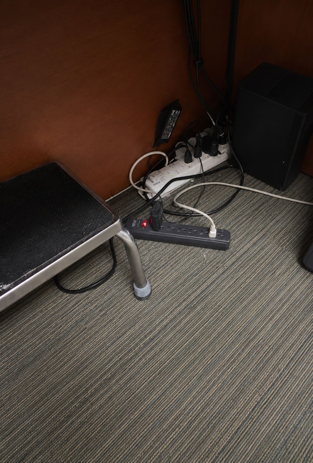 A grey power strip on carpet plugged into a white power strip behind it — two relocatable power taps in series under a desk